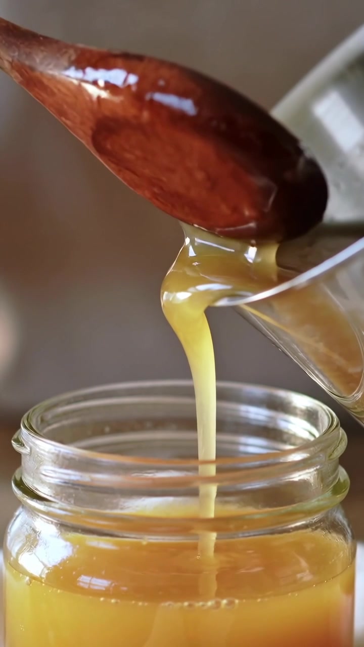 Video verticale: Mano che versa liquido dorato da un cucchiaio in un barattolo di vetro in cucina, inclinando il beccuccio.