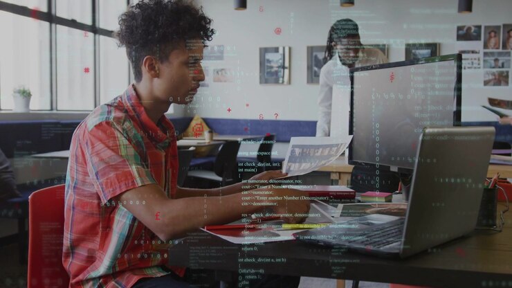 Seated man scanning printout at table, typing on laptop for business while colleagues passing