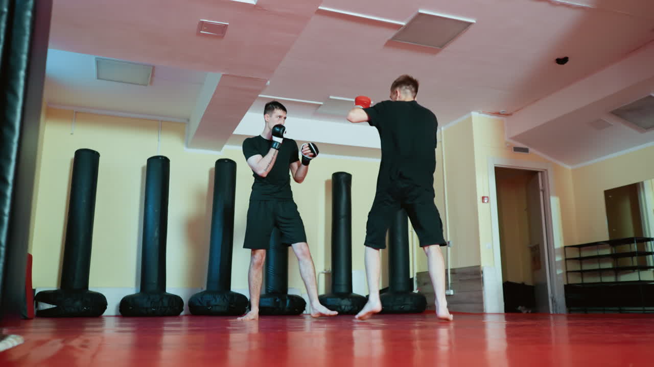 Athletes engaged in martial arts sparring session inside gym, wearing black training outfits and gloves, practicing combat sport techniques with defensive stances and kicks on red mat floor