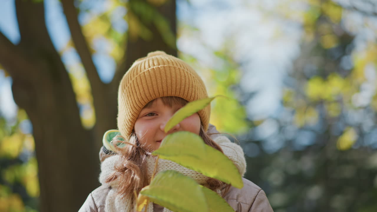 close up of little girl with warm smile holding yellow autumn leaf under tree canopy on leaf strewn park path during crisp fall day under cloudy sky capturing joyful moment