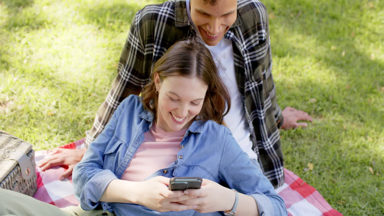 Happy diverse couple having picnic and using smartphone in sunny garden, in slow motion