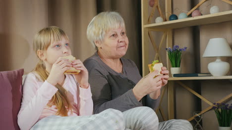 woman with granddaughter together watching tv and eating sandwiches