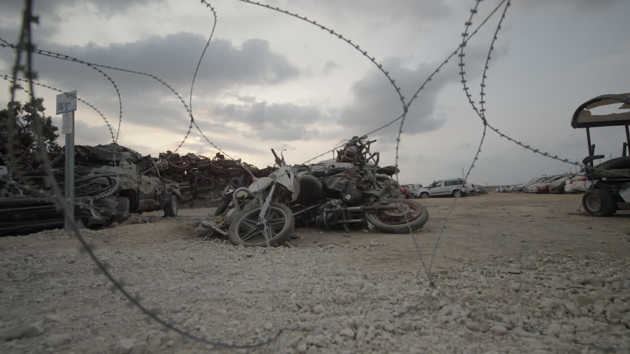 Wide shot of destroyed motorcycles looking through Barbed Wire at the vehicle lot memorial, Tkuma Israel after Hamas Attack