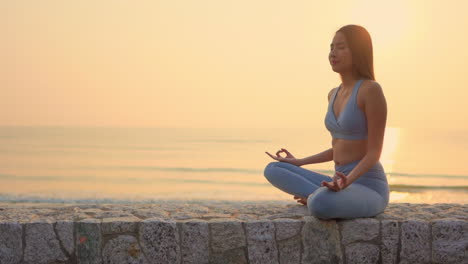 mujer asiática sentada en una pared junto al mar al atardecer en posición de loto meditando