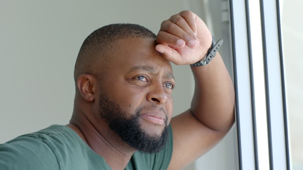 African American man seeing outside, standing at window in green T-shirt, raising hand calming gaze