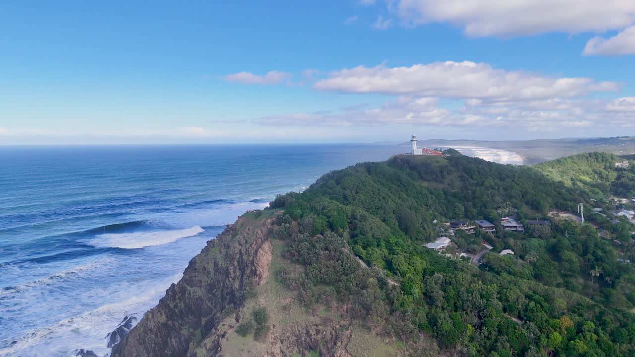 Drone footage captures Byron Bay lighthouse, lush greenery, and expansive coastline under clear skies with gentle waves