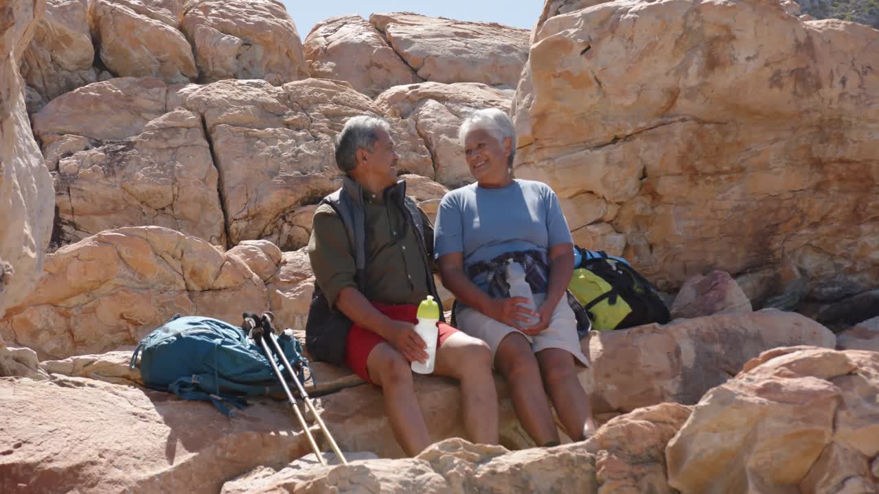 Happy senior biracial couple in mountains resting at sea, in slow motion