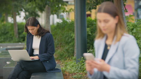 female office workers outdoors working on laptop and using mobile phone during break from the office