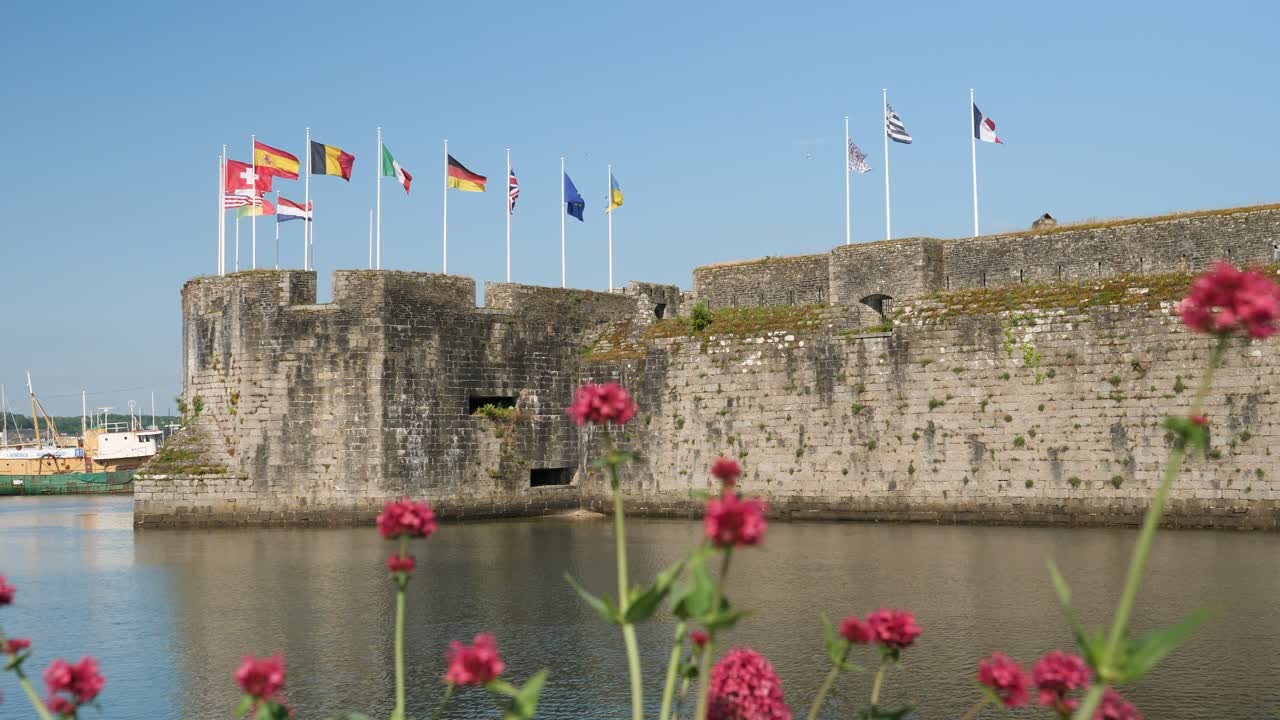 Scatto statico in una giornata di sole con cielo azzurro limpido, diverse bandiere europee sventolano sopra la città medievale fortificata di Concarneau, Bretagna, con fiori rosa in primo piano