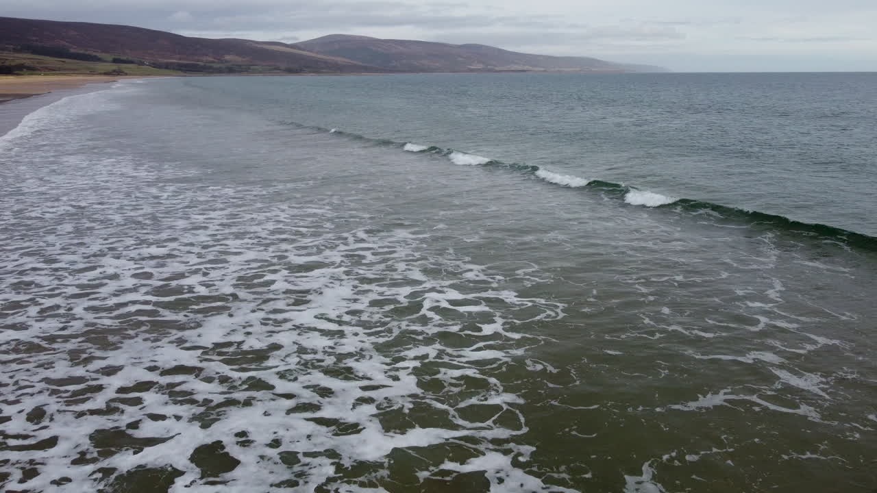 짙은 녹색 해안 파도가 울창한 산들을 배경으로 부서집니다.