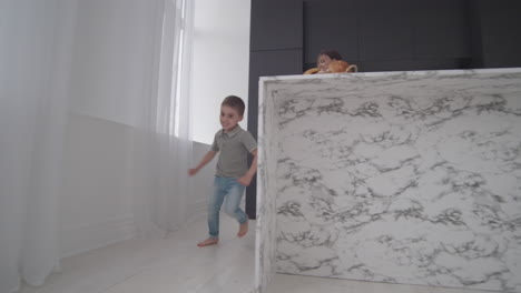 mother and baby son playing tag running through the house and laughing. son runs away from his mother and hides in the kitchen