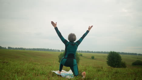 back view of woman in green and black suit outdoor in vast grassy field on yoga mat practicing low lunge pose, arms extended, under a cloudy sky with trees lined up in background