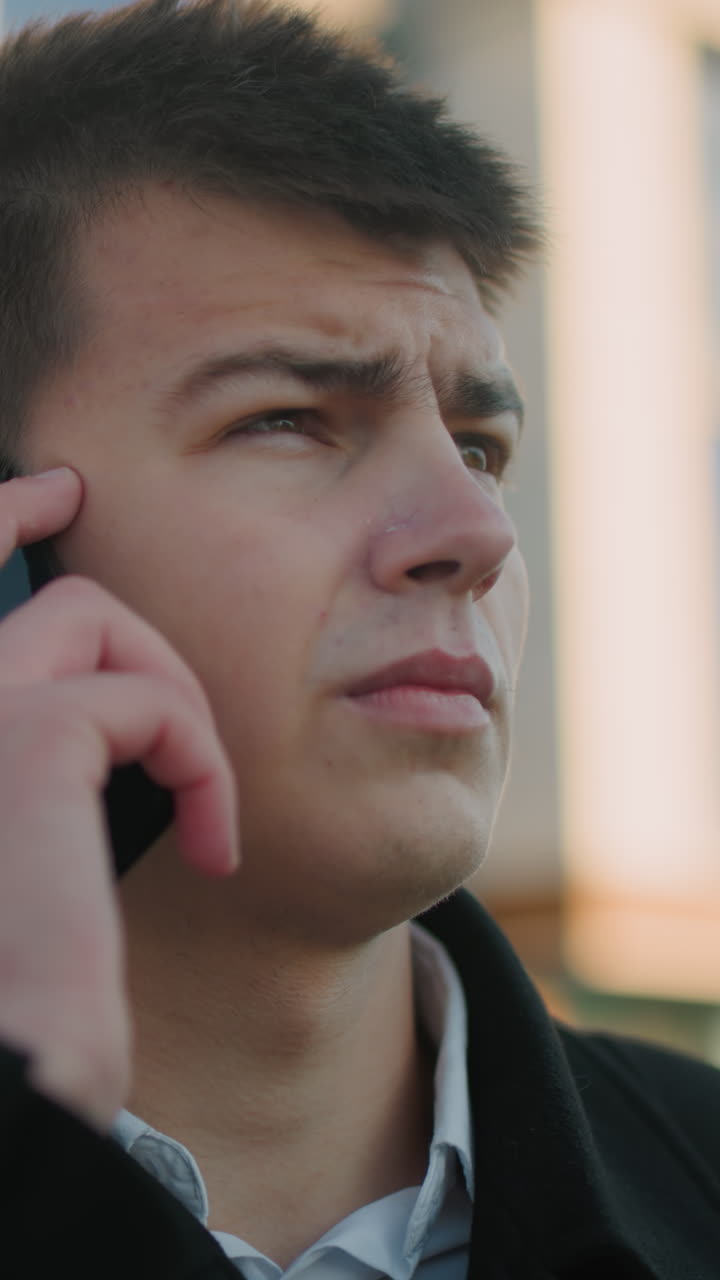 Business tycoon in black suit with white shirt answering phone call outdoors while nodding in agreement, background features modern glass building with soft light