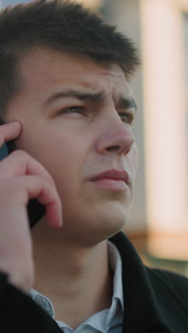 business tycoon in black suit with white shirt answering phone call outdoors while nodding in agreement, background features modern glass building with soft light