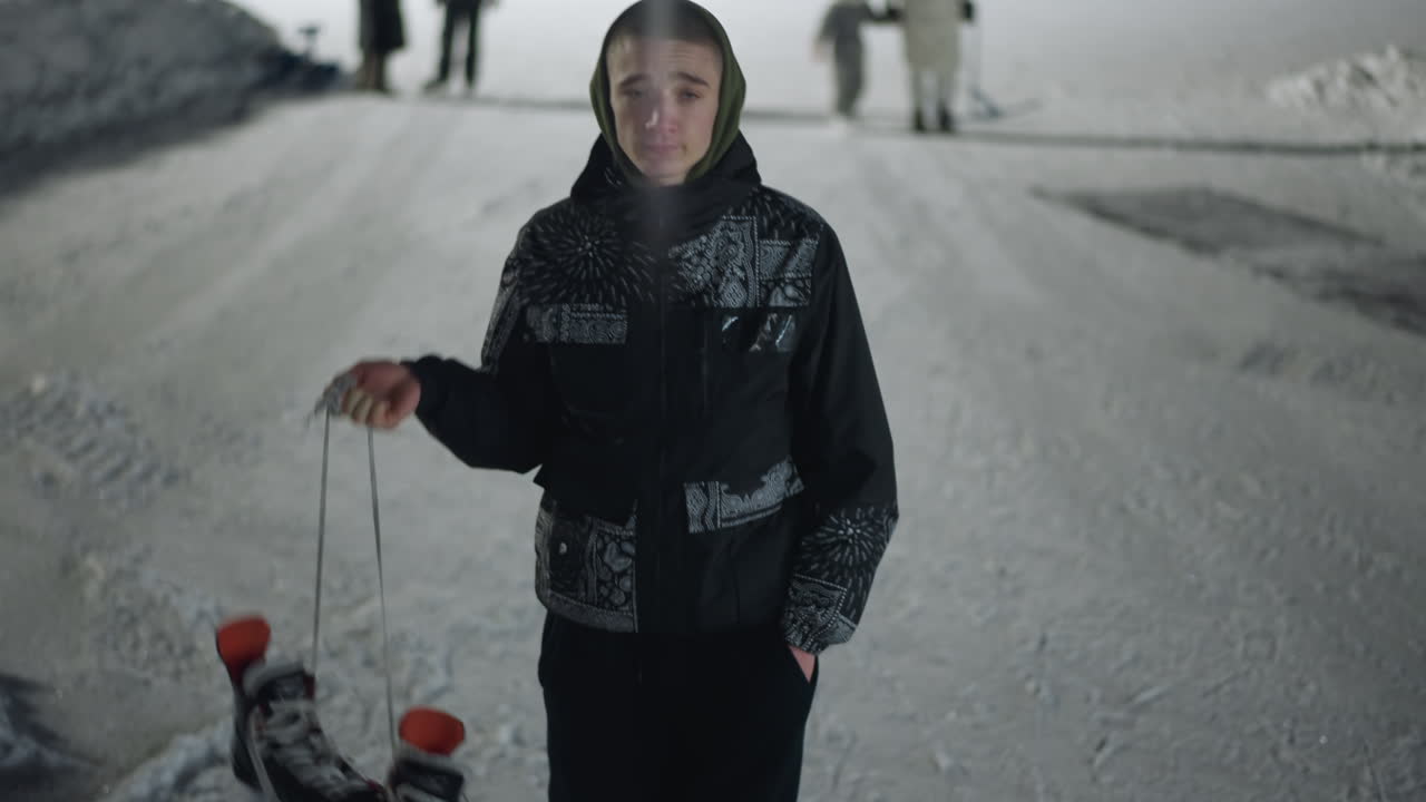 young skater in hooded jacket holding skate boot over shoulder walks across icy surface after night skating as people remain in background under soft light in cold winter environment