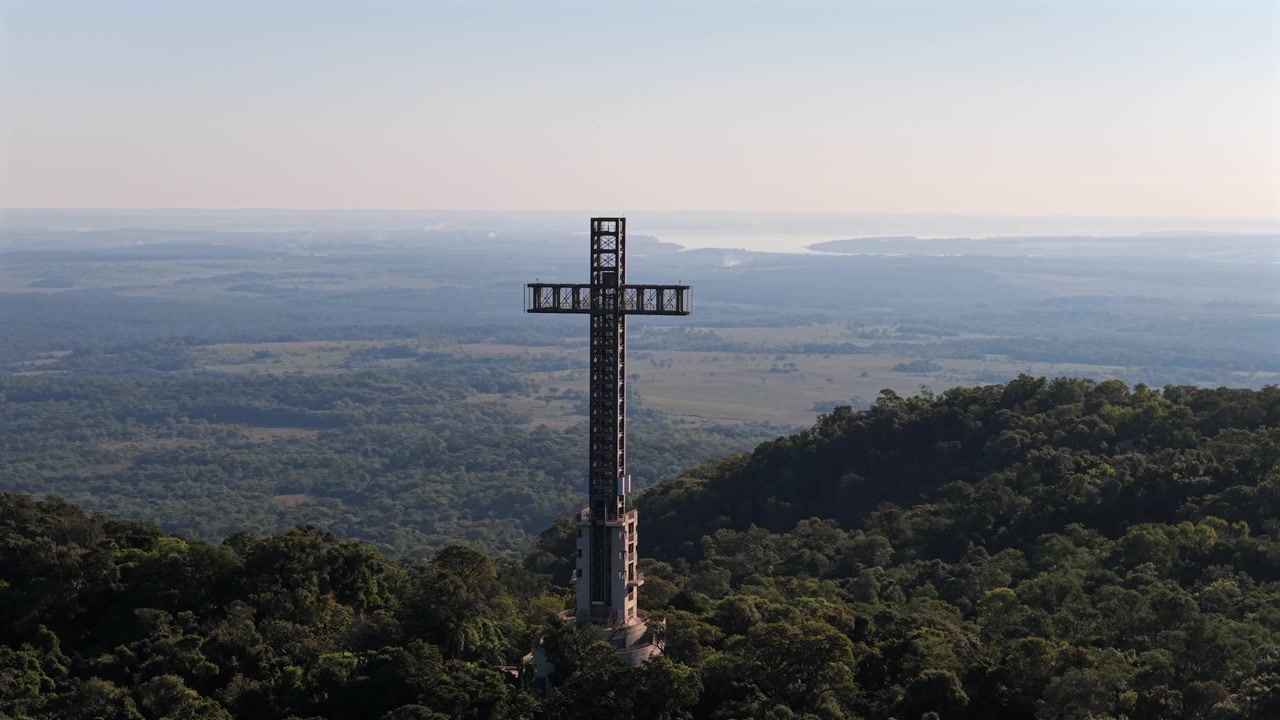 A cruz monumental ergue-se imponente em meio à vegetação exuberante, com o Rio Paraná ao fundo, em um dia ensolarado. Ideal para documentários de viagem ou reportagens culturais.