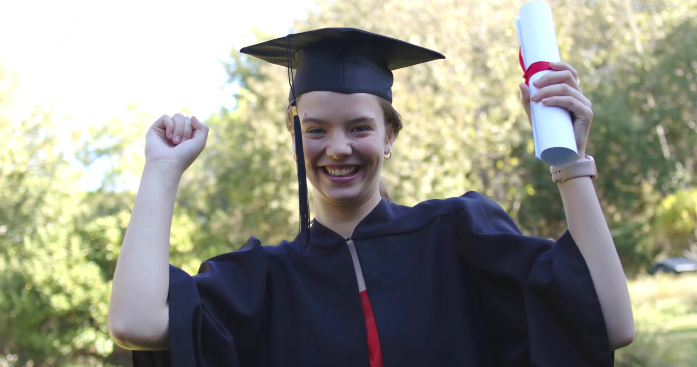 Giovane donna sui vent'anni che esulta con il diploma e alza il pugno dopo la cerimonia, in tocco e toga, in un parco soleggiato.