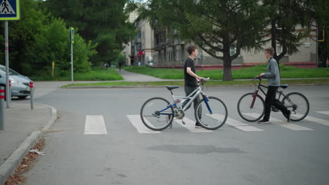 dos personas están cruzando un cruce de peatones marcado con pintura blanca en direcciones opuestas, uno está caminando con una bicicleta, y se ve un coche viniendo de la esquina, con el edificio en el fondo