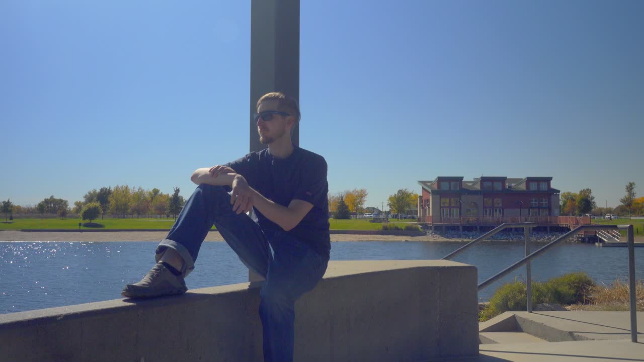 한 남자가 호수를 내려다보는  ⁇ 대 위에 앉아서  ⁇ 드폰을 꺼낸다.