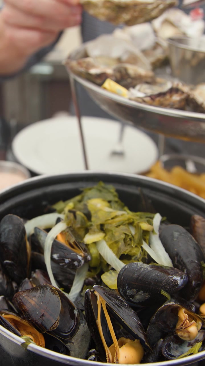 Diner serves oysters and mussels with shrimp, bread, and wine at outdoor coastal restaurant table
