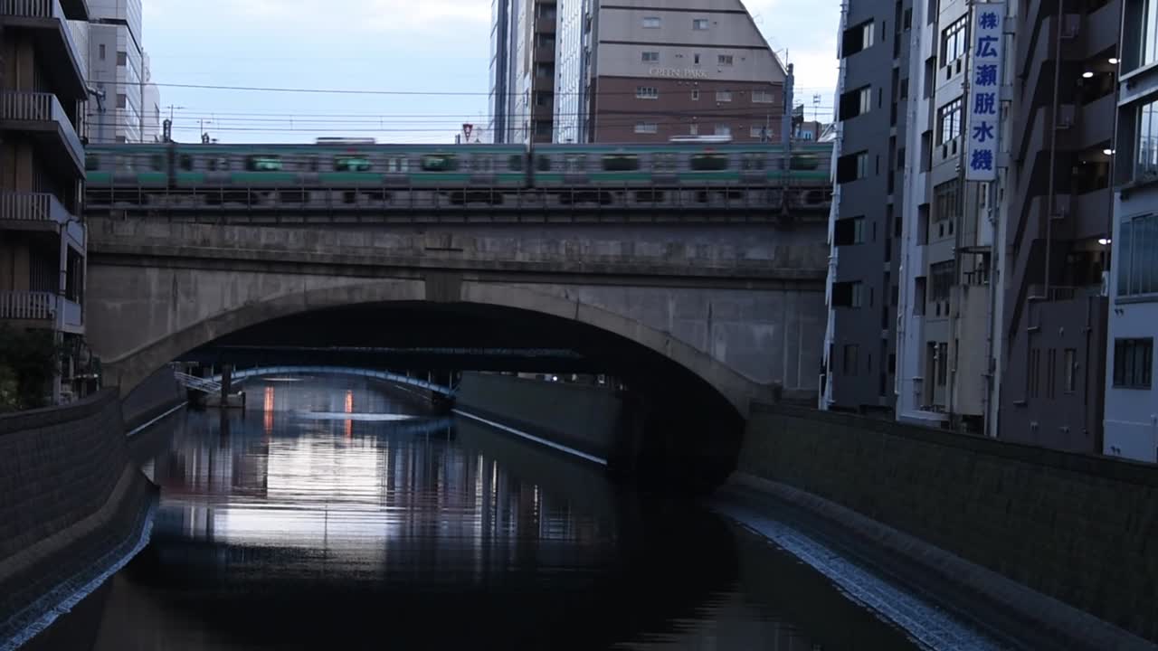 A train passes over a concrete bridge spanning a quiet urban canal at dusk in Tokyo