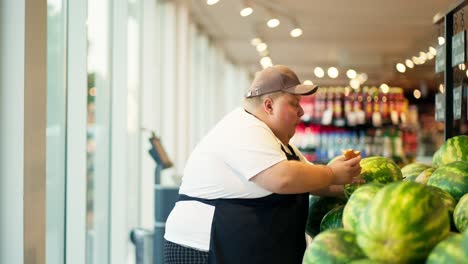 An-overweight-male-supermarket-worker,-a-man-in-a-white-T-shirt,-a-black-apron-and-a-gray-hat,-arranges-watermelons-and-checks-their-quality-on-the-supermarket-counter-while-eating-a-croissant