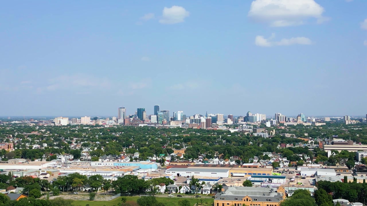 Ripresa statica con drone teleobiettivo strettamente inquadrata sugli edifici del centro di Winnipeg, con i quartieri circostanti e spazi verdi visibili sotto.