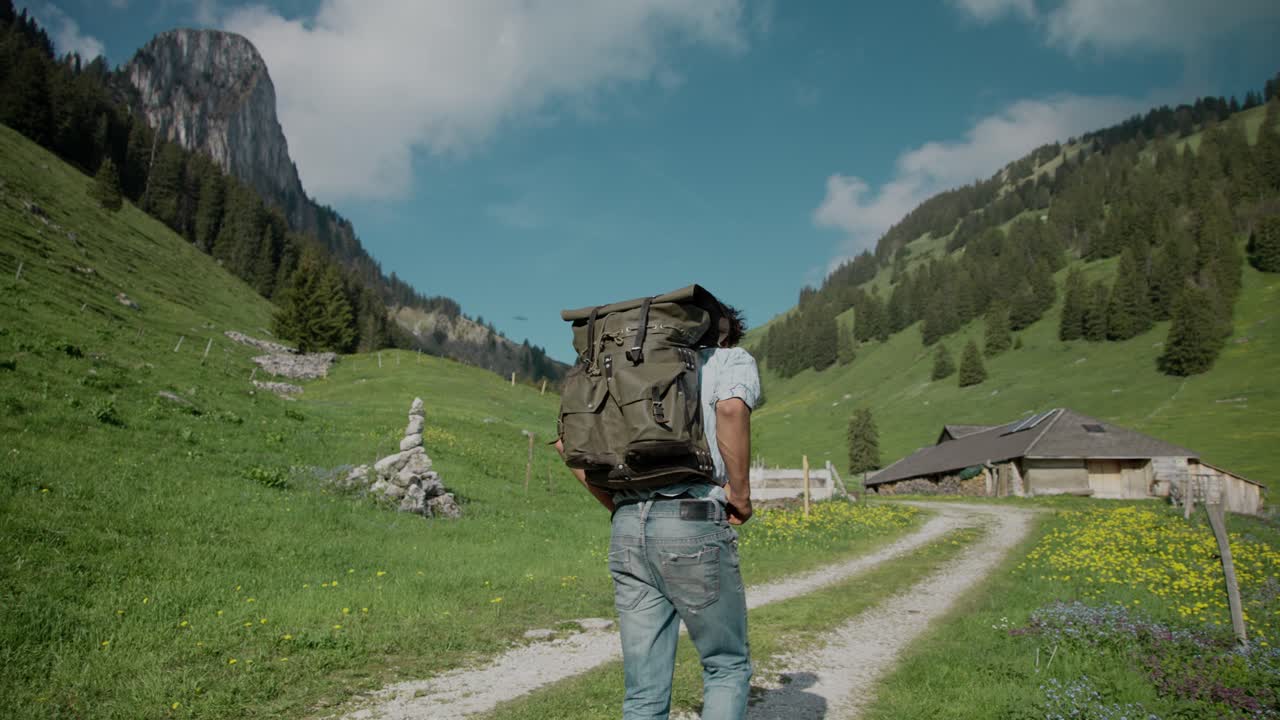 hombre milenario con ropa informal con mochila caminando por el sendero del valle de la montaña en cámara lenta, camino hacia una casa
