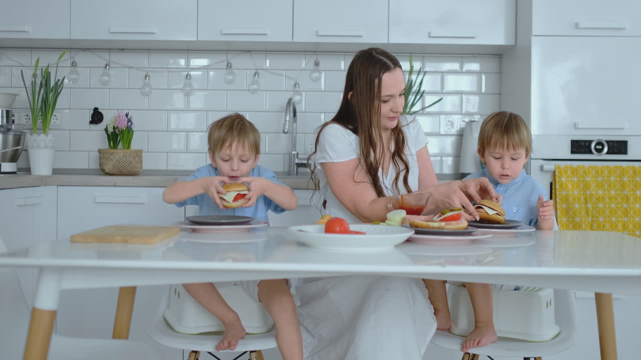 una familia feliz es una joven madre hermosa con un vestido blanco con dos hijos con camisas azules preparando una cocina blanca juntos cortando verduras y creando berger saludable para los niños.
