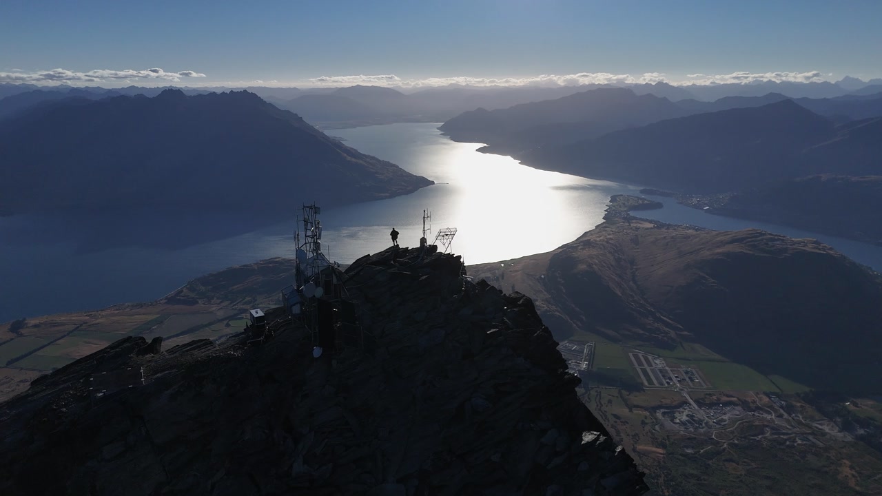 Aerial silhouette of man at comm tower on mtn peak above silvery lake