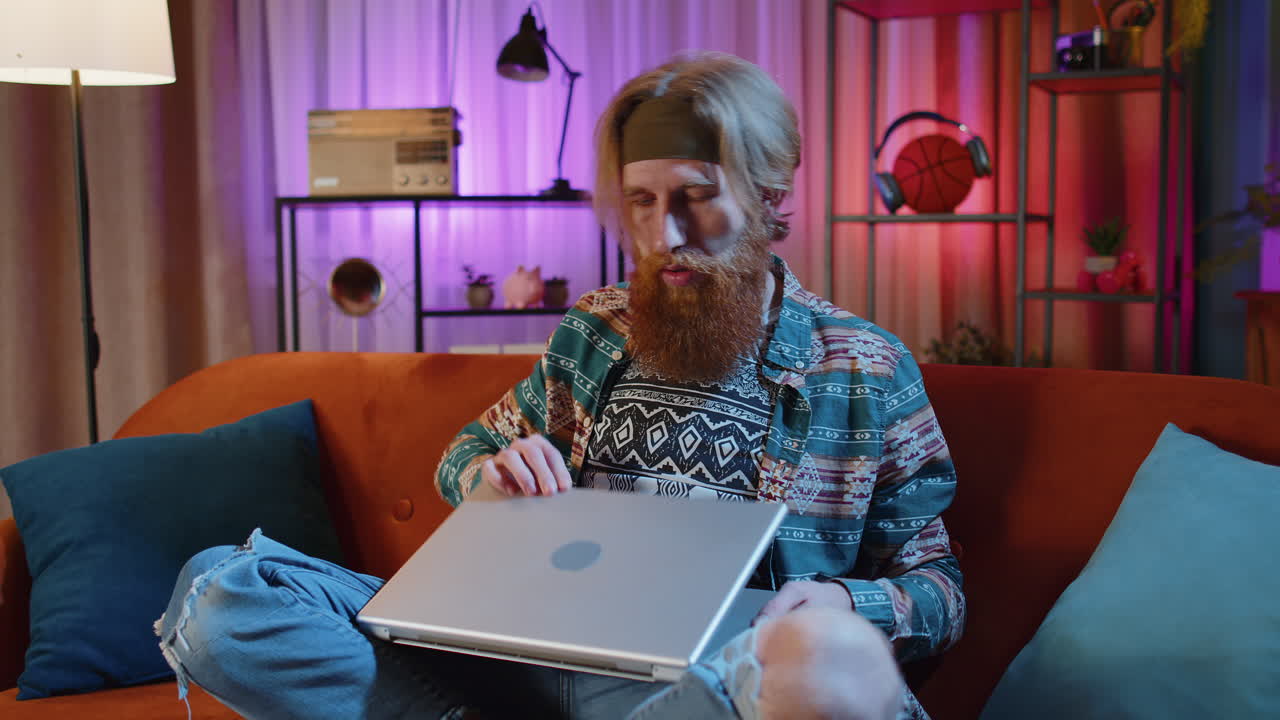 joven profesional sentado en el sofá cerrando la computadora portátil después de terminar el trabajo en la habitación de noche en casa