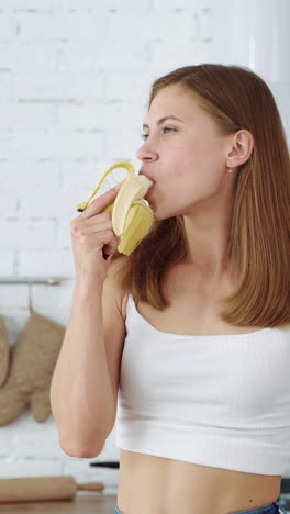 a young girl eats a banana on a white kitchen background. vertical video