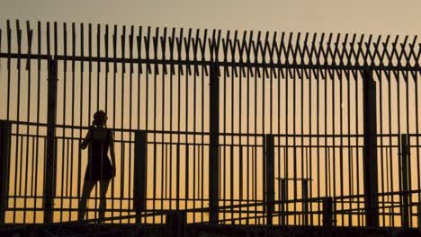 Silhouette-Einer-Jungen-Frau,-Die-Bei-Sonnenuntergang-Vor-Dem-Zaun-Spazieren-Geht