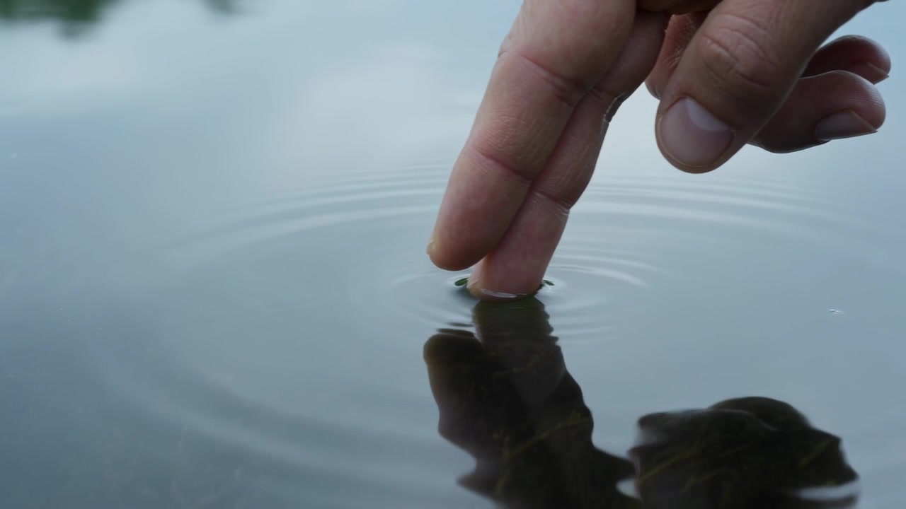 Polpastrello nudo che si avvicina e tocca la superficie di uno stagno, creando increspature e riflessi, con spazio per il testo.