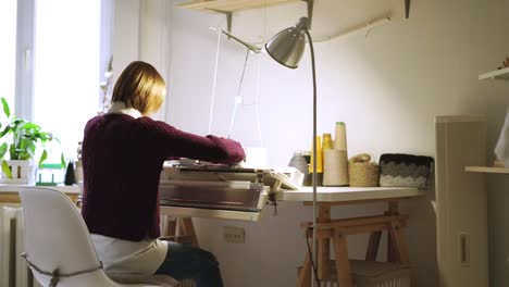 Young-woman-knitting-textile-on-weaving-machine-sitting-at-table-in-workshop