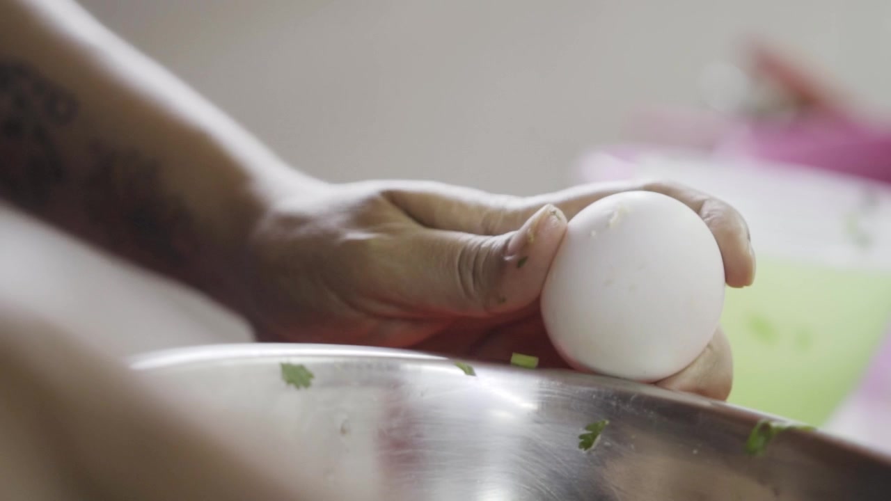 Closeup view of hand breaking egg above metal bowl with ingredients, step of adding egg to chicken meatball mixture, simple home cooking preparation in kitchen setting