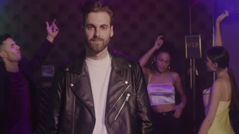 portrait of handsome bearded man in leather jacket smiling at camera at disco while his friends dancing behind him