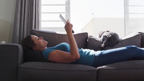 mujer caucásica acostada en el sofá con un perro usando una tableta digital