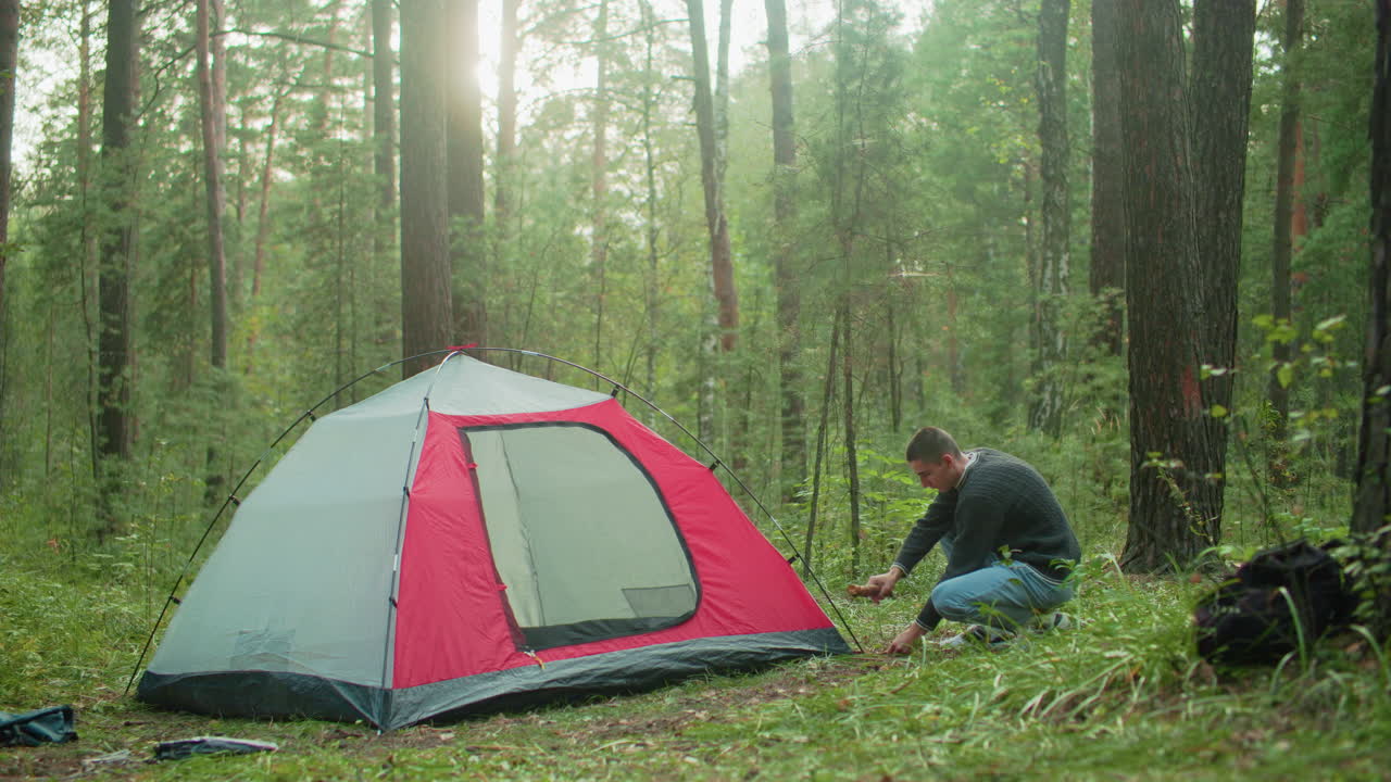 young man crouches in green forest beside red and gray tent using short wood stick to drive metal peg into ground with focused effort as sunlight filters through trees during outdoor