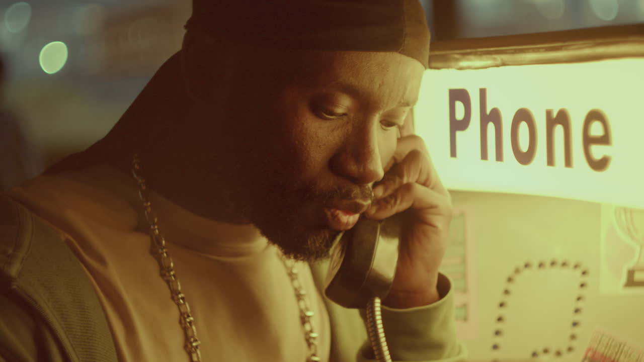 아프리카계 미국인 남자가 저녁에 거리의 공중전화에서 격렬한 대화를 나누고 있습니다.