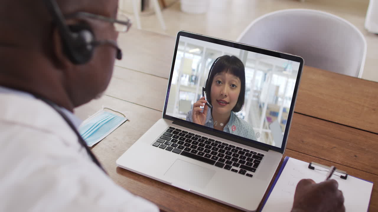 Ein afroamerikanischer Arzt mit Telefon-Headset macht sich Notizen während eines Videoanrufs am Laptop.