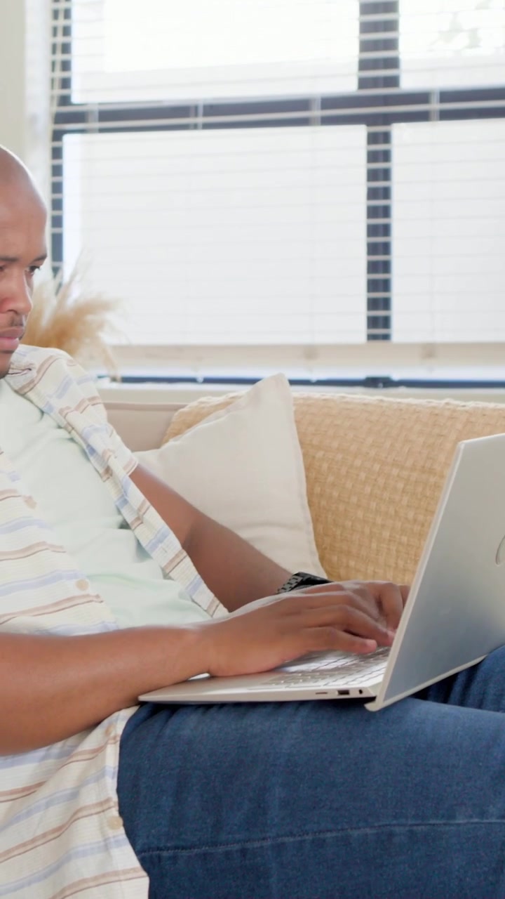 Vertical video: Scrolling man in striped top on couch placing phone down, typing on laptop to work
