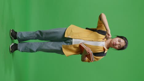 full body of asian woman student with a backpack and some books shaking head and showing thumbs down gesture while standing in the green screen background studio