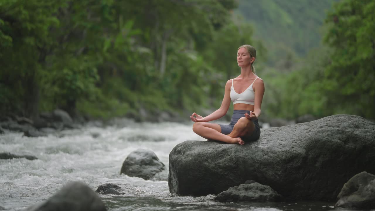 Attractive European blond female sitting on rock in lotus position, nature healing