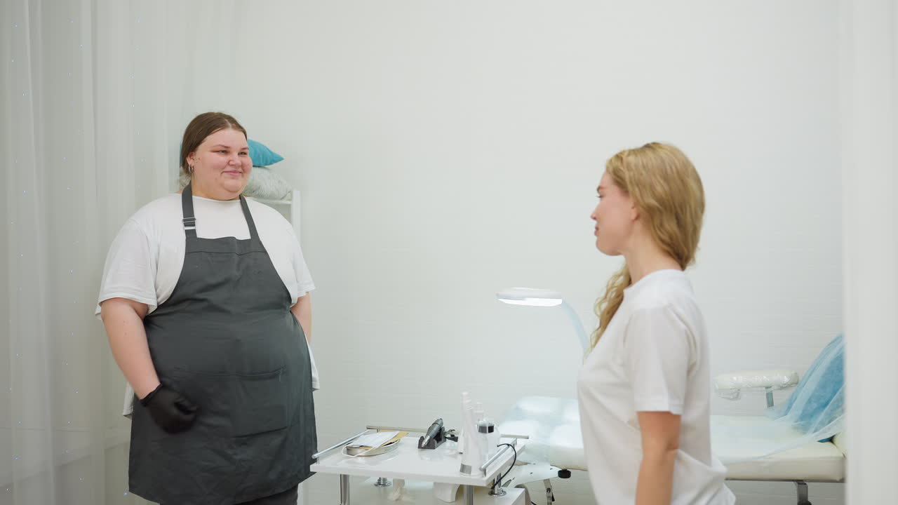 Beautician wearing apron and gloves smiles while welcoming client to treatment bed in bright, modern salon room with skincare tools arranged neatly on white table beside her