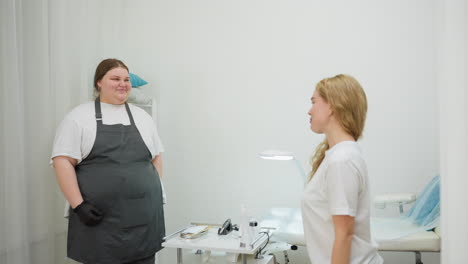 beautician wearing apron and gloves smiles while welcoming client to treatment bed in bright, modern salon room with skincare tools arranged neatly on white table beside her