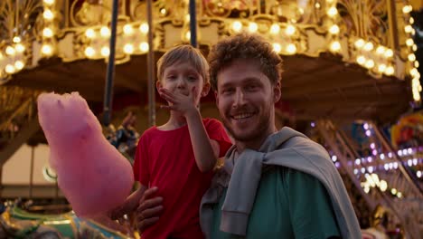 portrait of a happy blond boy in a red t-shirt and his dad eating pink cotton candy in the amusement park opposite against the backdrop of a beautiful bright attraction in yellow colors in the evening