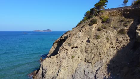 Paller-Des-Camp,-Felsnadelnadel,-Großes-Segelboot-In-Der-Ferne-Dramatische-Luftaufnahme-Flugsockel-Nach-Oben-Drohnenaufnahmen-Von-Aigües-Blanques-Strand-Ibiza-Sommertag-Juli-2022-Marnitz-4k-Film-Von-Oben