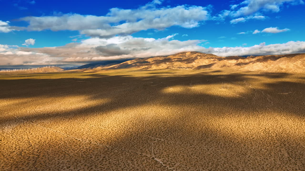 Dry deserted land with some spots of desert plants. Clouds float by the sky and throw shadows on the landscape. Top view.