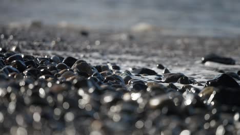 acercamiento extremo de las olas que se lavan en una playa pedregosa con tonos oscuros y marrones y reflejos del brillo en las rocas